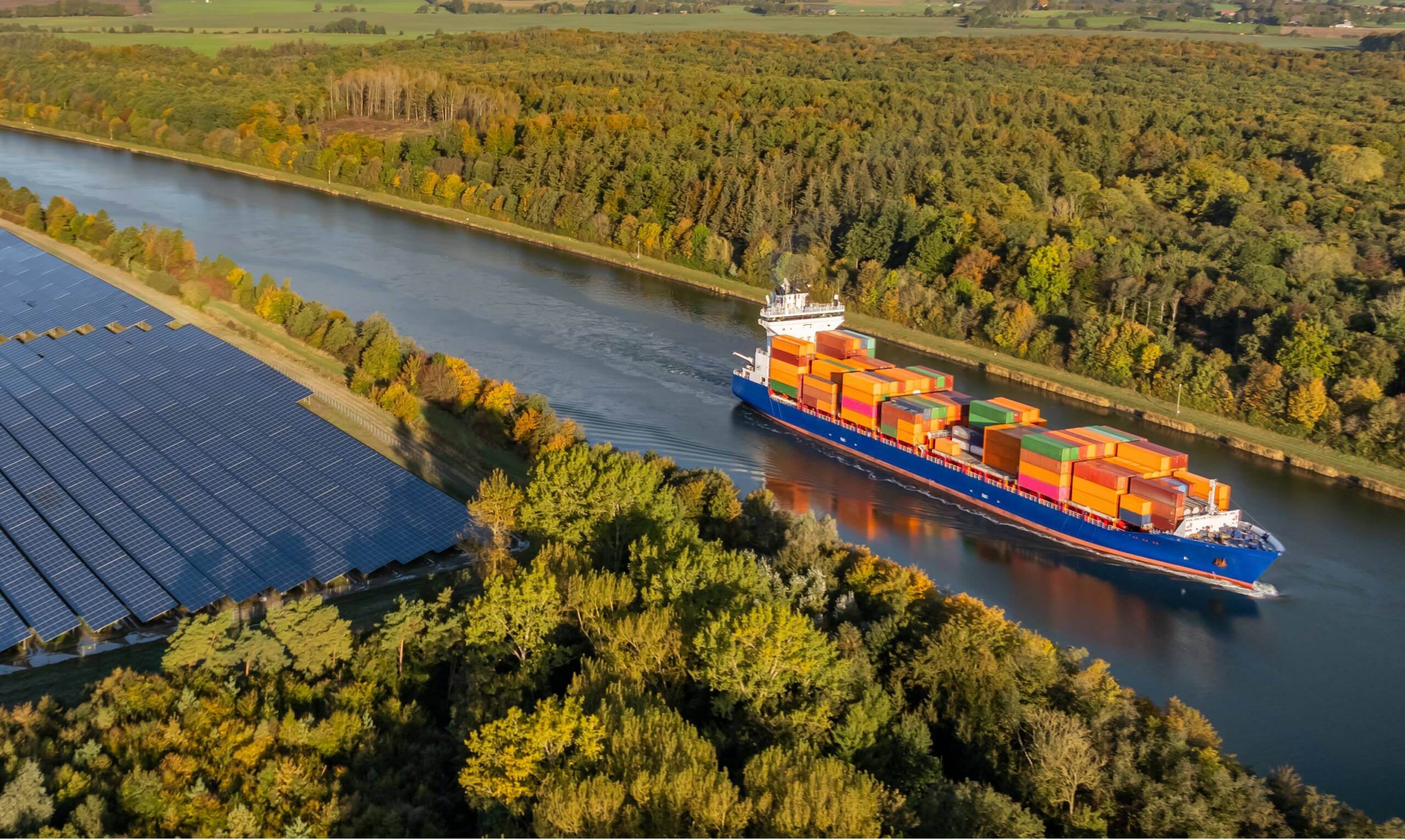 Vue aérienne d’une barge porte-conteneurs naviguant sur une voie fluviale en Europe, bordée de forêts et de panneaux solaires, illustrant le transport multimodal et durable (fleuve, rail, mer) pour une logistique décarbonée et une supply chain verte. - Yélé Consulting