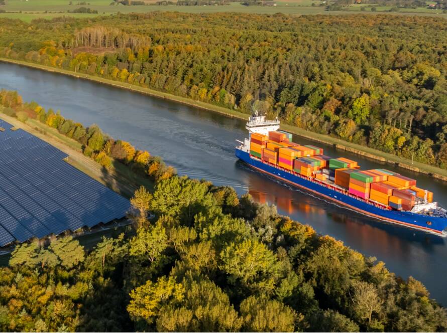 Vue aérienne d’une barge porte-conteneurs naviguant sur une voie fluviale en Europe, bordée de forêts et de panneaux solaires, illustrant le transport multimodal et durable (fleuve, rail, mer) pour une logistique décarbonée et une supply chain verte. - Yélé Consulting